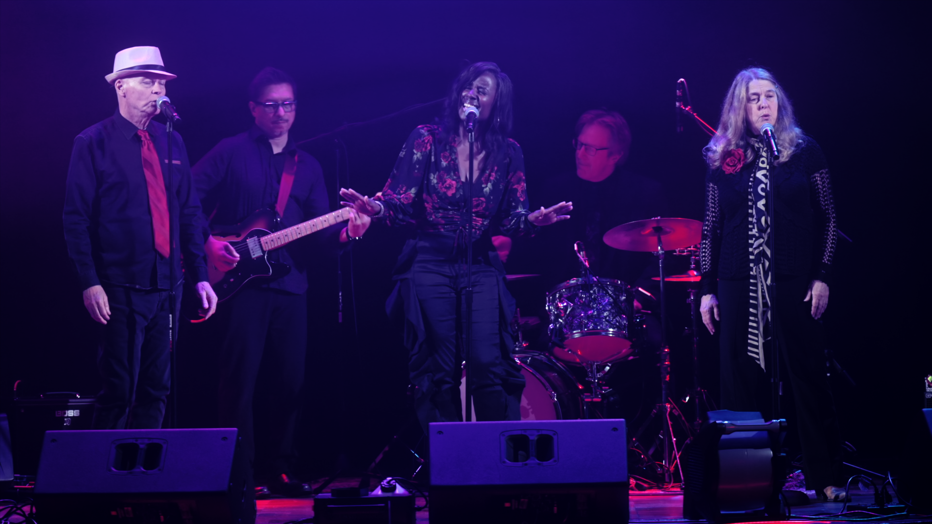 Five-member band performing on a purple-lit stage: two female singers at microphones center and right, with guitarist, bassist, and drummer in back.